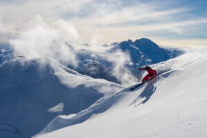 Skier skiing through powder with snow covered mountains behind them